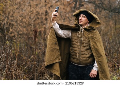 Portrait of survivalist male wearing tent raincoat searching for reception signal, making video call or recording video message on smartphone, surrounded by forest at outdoors in overcast cold day. - Powered by Shutterstock