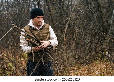 Portrait Of Survivalist Male Wearing Warm Clothes Collecting Dry Deadwood For Fire In Forest On Overcast Cold Day. Concept Of Exploration, Travel And Adventure In Nature.