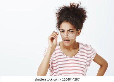 Portrait of suspicious intense funny woman with afro haircut, taking off glasses, bending towards camera and frowning, having doubts and gazing with disbelief at camera while standing over gray wall