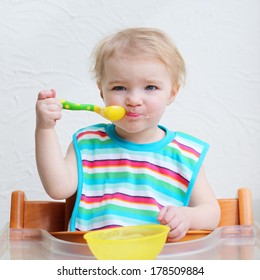 Portrait Of Sweet Blonde Toddler Girl Wearing Colorful Bib Eating Porridge With Spoon Sitting Indoors In High Chair With Table Against A White Wall