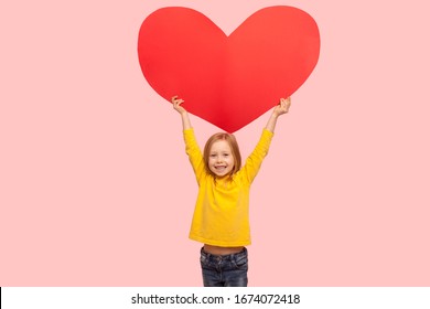 Portrait Of Sweet Cheerful Little Girl Raising Large Red Heart Symbol Over Head And Smiling To Camera, Congratulating On Mother's Day, Saying I Love You. Indoor Studio Shot Isolated On Pink Background