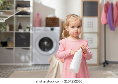 Portrait Sweet Little Girl Plays In The Laundry Room Bathroom Next To The Laundry Basket With Clothes. Child Holds Bottle For Liquid, Soap Opens Cap Looks In, Curiosity Learning About The World.