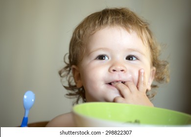 Portrait Of Sweet Small Smiling Baby Boy With Blonde Curly Hair And Round Cheecks Eating From Green Plate Holding Spoon And Lick Fingers Closeup, Horizontal Picture