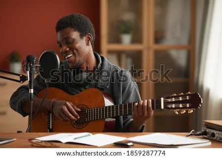 Portrait of talented African-American man singing to microphone and playing guitar while recording music in studio, copy space