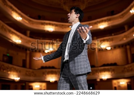 Portrait of talented attractive singer in elegant classic outfit, singing hit with open mouth in microphone gesture with hand standing on stage in theater. Handsome caucasian guy during performance