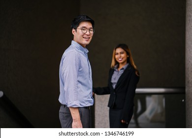 Portrait Of A Tall Asian Man Talking To A Beautiful And Young Asian Businesswoman In A Business Suit Pulling Her Business Suitcase. The Man Is Looking Back Over His Shoulder As If Someone Called Him.