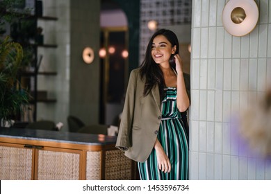 Portrait of a tall, slim, elegant and beautiful Indian Asian woman in an elegant jumper with a green jacket draped over her shoulders. She is smiling as she stands in a well-appointed home or room.