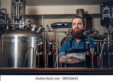 Portrait of tattooed, bearded hipster male manufacturer presenting beer in the microbrewery.