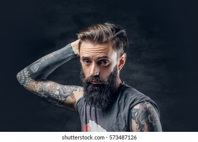 Portrait of a tattooed, bearded male in a t shirt touches his head over a dark background.