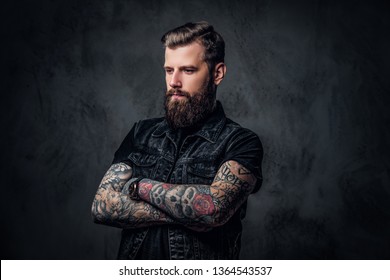 Portrait of a tattooed man with beard and hairstyle posing with his arms crossed. Studio photo against a dark wall