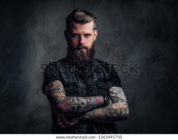 Portrait of a\
tattooed man with beard and hairstyle posing with his arms crossed.\
Studio photo against a dark\
wall