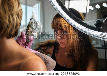 Portrait of tattoo-master makes a tattoo to a client. View from the female's shoulder. Concept of world tattoo day.