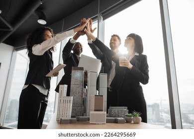 Portrait of team of positive successful multiethnic corporate coworkers at the office meeting, standing near table with 3d city model and giving high five as a sign fruitful collaboration. - Shutterstock ID 2264295669