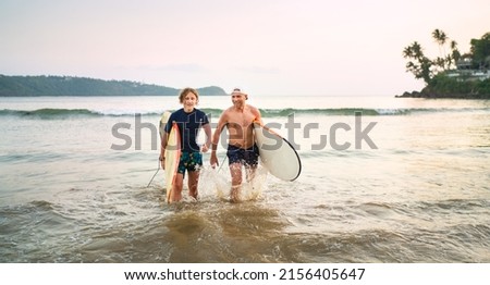 Portrait of a teen boy with his father with surfboards after surfing. They are smiling and walking out of the water. Family active vacation or active sporty people concept.