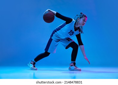 Portrait Of Teen Girl, Basketball Player In Motion, Dribbling Ball Isolated Over Blue Studio Background In Neon. Concept Of Sport, Active Lifestyle, Health, Team Game, Hobby And Ad