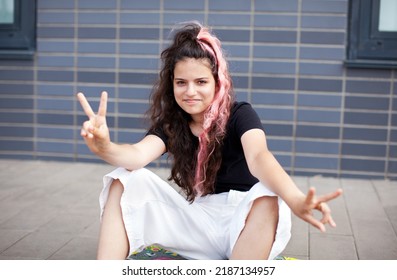 Portrait Teen Girl With Long Dark Hair Dyed Pink, Looking In Camera And Shows Peace Sign By Hand On Grey Background