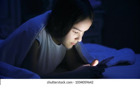 Portrait Of Teen Girl Lying In Bed At Night And Using Smart Phone