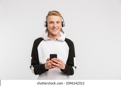 Portrait Of Teenage Boy 16-18 Years Old Wearing Hoodie Smiling And Listening To Music On Smartphone Via Wireless Headphones Isolated Over White Background