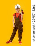 Portrait of teenage boy in work jumpsuit and protective helmet holds drill. Child is like worker builder on yellow background