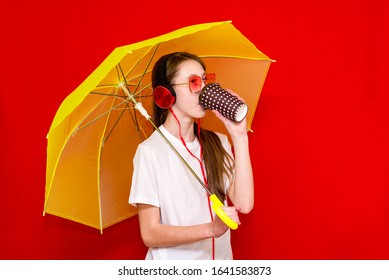 Portrait Of Teenage Caucasian Girl Listens To Music In Headphones With Yellow Umbrella Sunglasses Drink Coffee In Red Studio Background Wearing White T Shirt