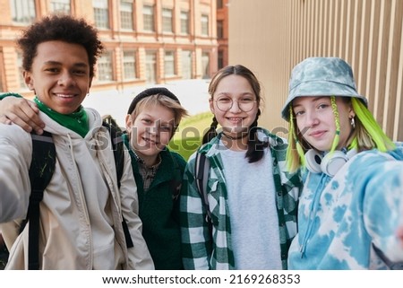 Portrait of teenage friends smiling at camera while making selfie portrait on smartphone standing outdoors with school building in background