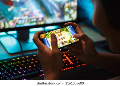 Portrait of teenage gamer boy playing video games on smartphone and computer in dark room wearing headphones and using backlit colorful keyboard