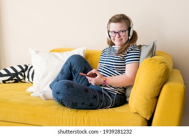 Portrait Of Teenage Girl With Down Syndrome Listening To Music In Headphones Sitting On Sofa At Home. Audio Development For Children With Disabilities, Inclusive Education