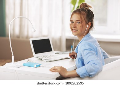 Portrait of teenage girl wearing headphones sitting at table smiling looking at camera doing homework using laptop, online education, distance learning