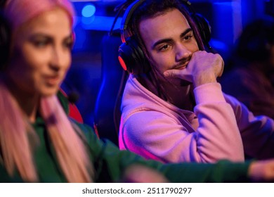 Portrait of a teenage professional gamer with dreadlocks and headset sitting in neon lighted internet cafe with his female friend and looking at her monitor. A gamer is helping his friend with game. - Powered by Shutterstock