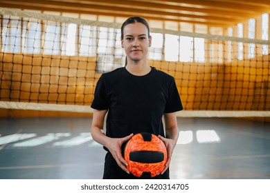 Portrait of a teenage professional sportswoman standing on volleyball court with a ball in her hands and looking at the camera. A fit teenage female volleyball player standing on court with ball. - Powered by Shutterstock