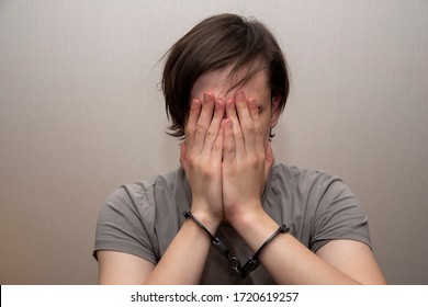 Portrait Of A Teenager In Handcuffs Covering His Face With His Hands On A Gray Background, Medium Plan. Juvenile Delinquent, Criminal Liability Of Minors. Members Of Youth Criminal Groups And Gangs.