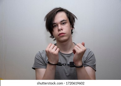 Portrait Of A Teenager In Handcuffs On A Gray Background, Medium Plan. Juvenile Delinquent, Criminal Liability Of Minors. Members Of Youth Criminal Groups And Gangs.