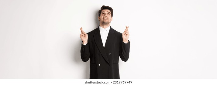 Portrait of tensed and worried handsome businessman, crossing fingers and looking up, begging god, making a wish, standing against white background in black suit. - Powered by Shutterstock