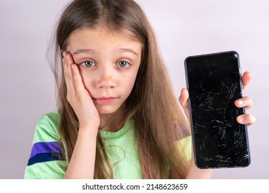 Portrait Of Terrified Child With Damaged Phone. Upset Little Girl Breaks Screen Of Mobile Phone. Kid Girl Holding Broken Display. Worried Child Accidentally Destroyed Smartphone