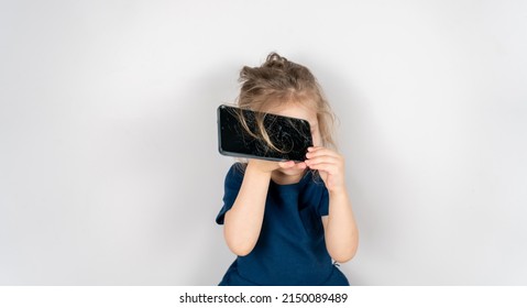 Portrait Of Terrified Child With Damaged Phone. Upset Little Girl Breaks Screen Of Mobile Phone. Kid Girl Holding Broken Display. Worried Child Accidentally Destroyed Smartphone