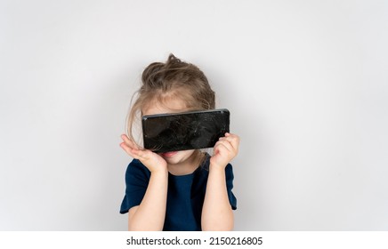 Portrait Of Terrified Child With Damaged Phone. Upset Little Girl Breaks Screen Of Mobile Phone. Kid Girl Holding Broken Display. Worried Child Accidentally Destroyed Smartphone