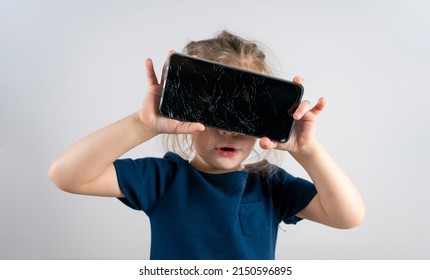 Portrait Of Terrified Child With Damaged Phone. Upset Little Girl Breaks Screen Of Mobile Phone. Kid Girl Holding Broken Display. Worried Child Accidentally Destroyed Smartphone