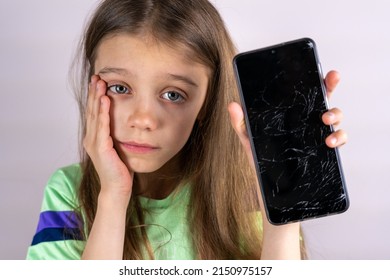 Portrait Of Terrified Child With Damaged Phone. Upset Little Girl Breaks Screen Of Mobile Phone. Kid Girl Holding Broken Display. Worried Child Accidentally Destroyed Smartphone