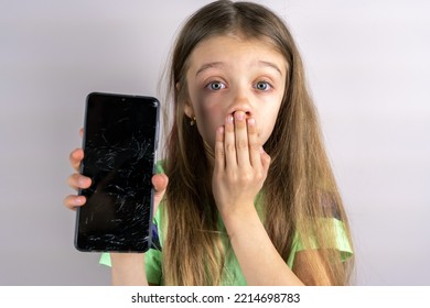 Portrait Of Terrified Child With Damaged Phone. Upset Little Girl Breaks Screen Of Mobile Phone. Kid Girl Holding Broken Display. Worried Child Accidentally Destroyed Smartphone