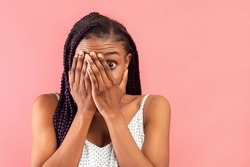 Portrait Of Terrified Young Black Lady In Dress Covering Her Face With Hands And Peeking Through Fingers Over Pink Studio Background, Copy Space. Phobia, Fear Concept