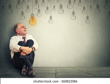 Portrait Thinking Elderly Man In Glasses Sitting On A Floor Looking Up With Light Idea Bulb Above Head Isolated On Gray Wall Background
