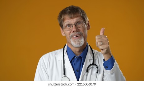 Portrait Of Thinking Pondering Doctor Man In Medical Coat Having Idea Moment Giving Thumbs Up On Solid Yellow Studio Background. Smiling Happy Doc Showing Eureka