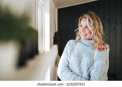 Portrait of thinking smiling woman forty year with blonde curly hair in grey knitted sweater near window at house - Shutterstock ID 2232507421