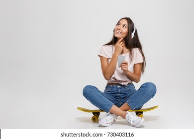 Portrait of thinking young woman in white headphone holding mobile phone, sitting on skateboard, looking up, isolated over white background - Powered by Shutterstock