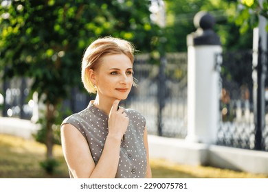Portrait of thoughtful 40 years old woman standing outdoors among growing green trees in alley and touching chin. Middle aged lady photo on sunny day. Selective focus, blurred urban background. - Shutterstock ID 2290075781
