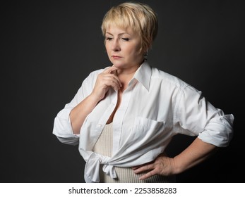 Portrait of a thoughtful adult woman with short blonde hair in a stylish white shirt . Photo studio, dark background. - Shutterstock ID 2253858283
