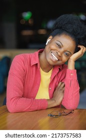 Portrait Of A Thoughtful African American Woman In A Pink Shirt And Glasses. A Happy Woman Is Relaxing In A Public Place And Posing. A Successful Confident Millennial Woman Smiles. A Student Gets An