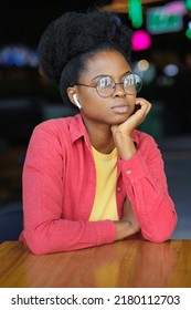 Portrait Of A Thoughtful African American Woman In A Pink Shirt And Glasses. A Happy Woman Is Relaxing In A Public Place And Posing. Selective Focus, Successful Confident Millennial Woman Smiles. A