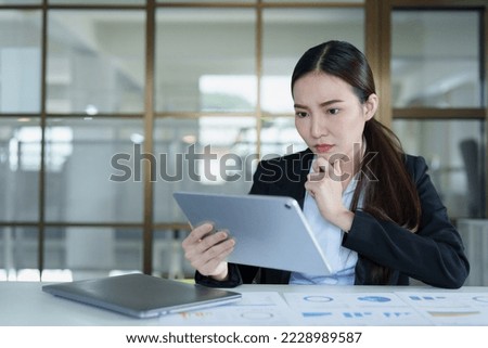 Portrait of a thoughtful Asian businesswoman looking at financial statements and making marketing plans using a tablet computer on her desk.