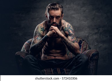 Portrait of a thoughtful bearded male with tattooed naked torso and crossed arms, sits on a chair over dark grey background.
